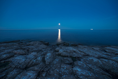 Reflected moonlight on a calm sea, viewed across a shoreline of mottled, jagged rock. Blue night and a glowing horizon; perhaps a distant ship.