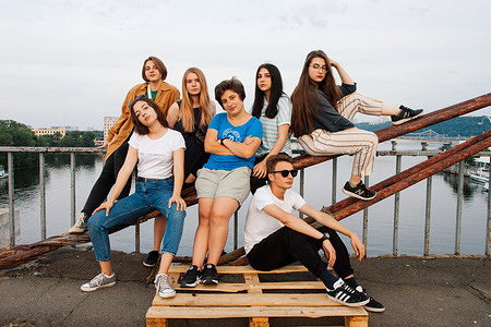 Group of young friends in cool pose, standing and sitting on pallet and metal bars, in front of railings by an urban waterfront.