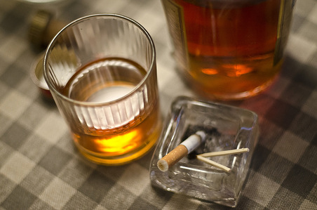 Close-up of a glass half filled with alcohol next to a cigarette in an ashtray.